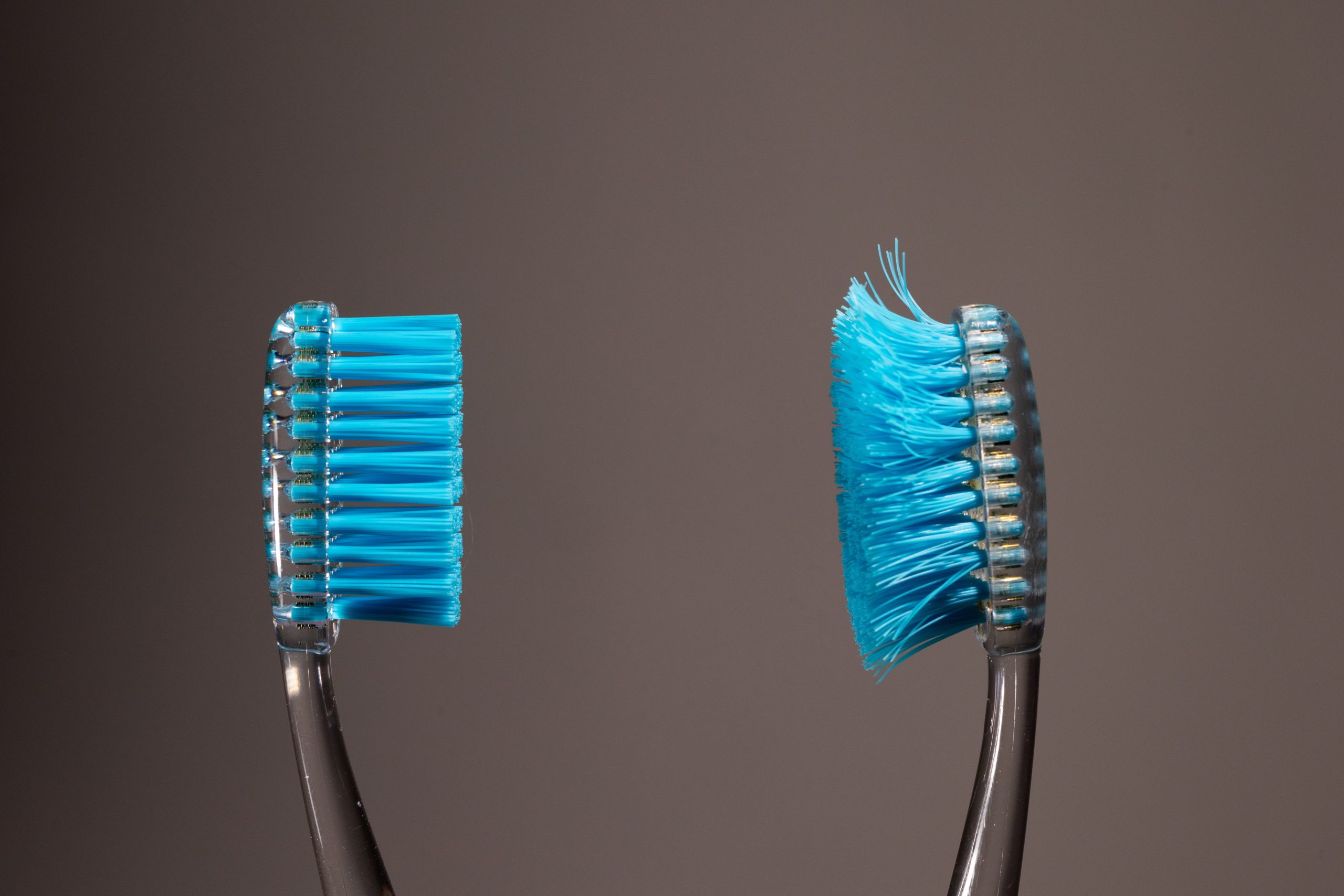 A side-by-side comparison of a brand new blue toothbrush next to a worn-out, frayed blue toothbrush on a dark background.